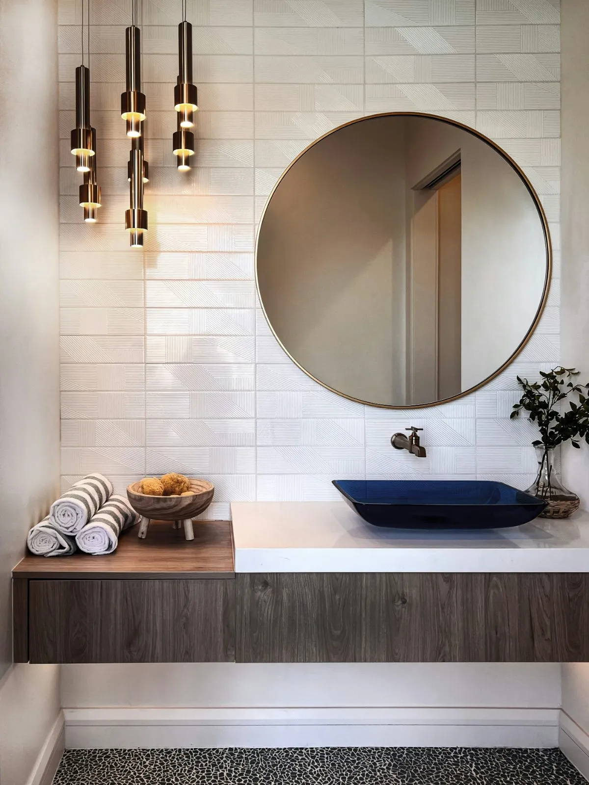 Textured white ceramic wall tile behind a floating vanity with a round mirror and brass pendants